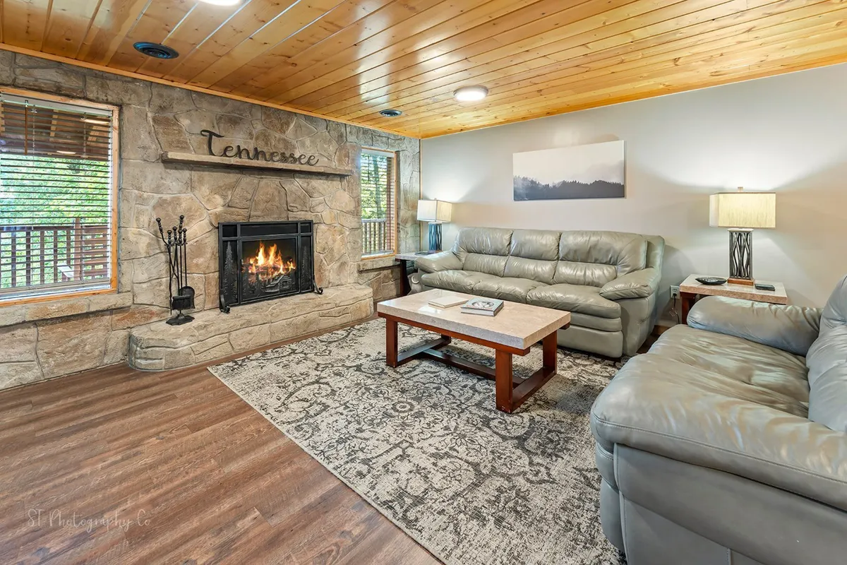 The image features a warm, inviting living room with a wood-burning fireplace as the focal point. Plush sofas and a coffee table are arranged around the hearth, and tasteful artwork adorns the walls. The setting is cozy and intimate, evoking the classic charm of a cabin retreat.
