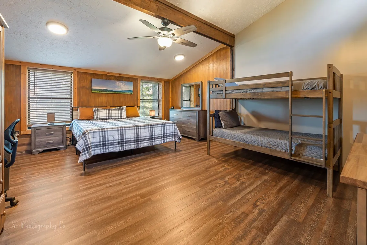 A spacious bedroom filled with natural light, featuring a large bed with plaid bedding, a cozy bunk bed tucked into the corner, wooden furniture, and a large window overlooking the outdoors. The inviting arrangement and warm tones create a welcoming retreat for families or groups, with space for both adults and children.