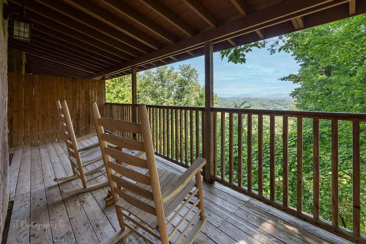 A private back deck featuring classic wooden rocking chairs, nestled against a sturdy wooden railing. The deck overlooks lush greenery and offers an expansive mountain view in the distance. The scene evokes tranquility with its soft natural light, suggesting an ideal spot for sunrise coffee or sunset conversations while soaking in the fresh mountain air.