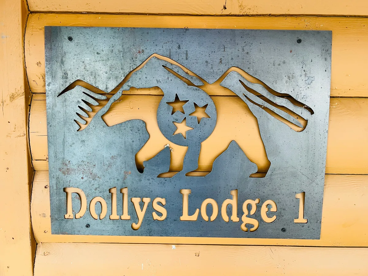A rustic metal sign featuring a bear and mountain motif and the text 'Dollys Lodge 1.' The sign welcomes guests at the lodge's entrance, evoking a sense of place and adventure while hinting at the wildlife and mountain scenery nearby.
