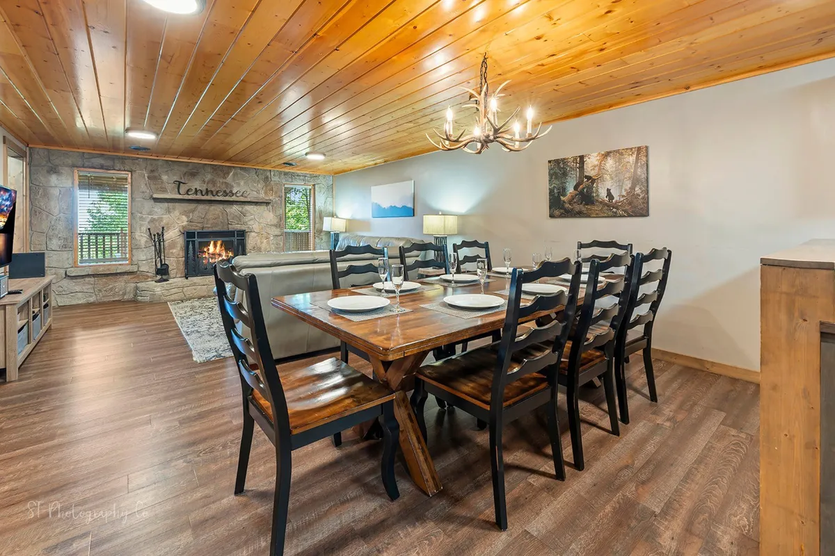 A spacious dining area with a rustic wooden table sits adjacent to a charming fireplace. A chandelier adds a touch of elegance, and large windows let in soft, natural light, hinting at scenic mountain views beyond. The overall mood is comfortable and welcoming—ideal for sharing meals or quiet moments.