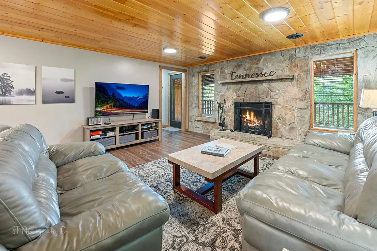 A comfortable living area centered on a stone wood-burning fireplace, with plush leather sofas and a coffee table—ideal for relaxing, board games, or storytelling. The abundance of natural light from the windows and the TV nearby help illustrate the entertainment options described in the section.