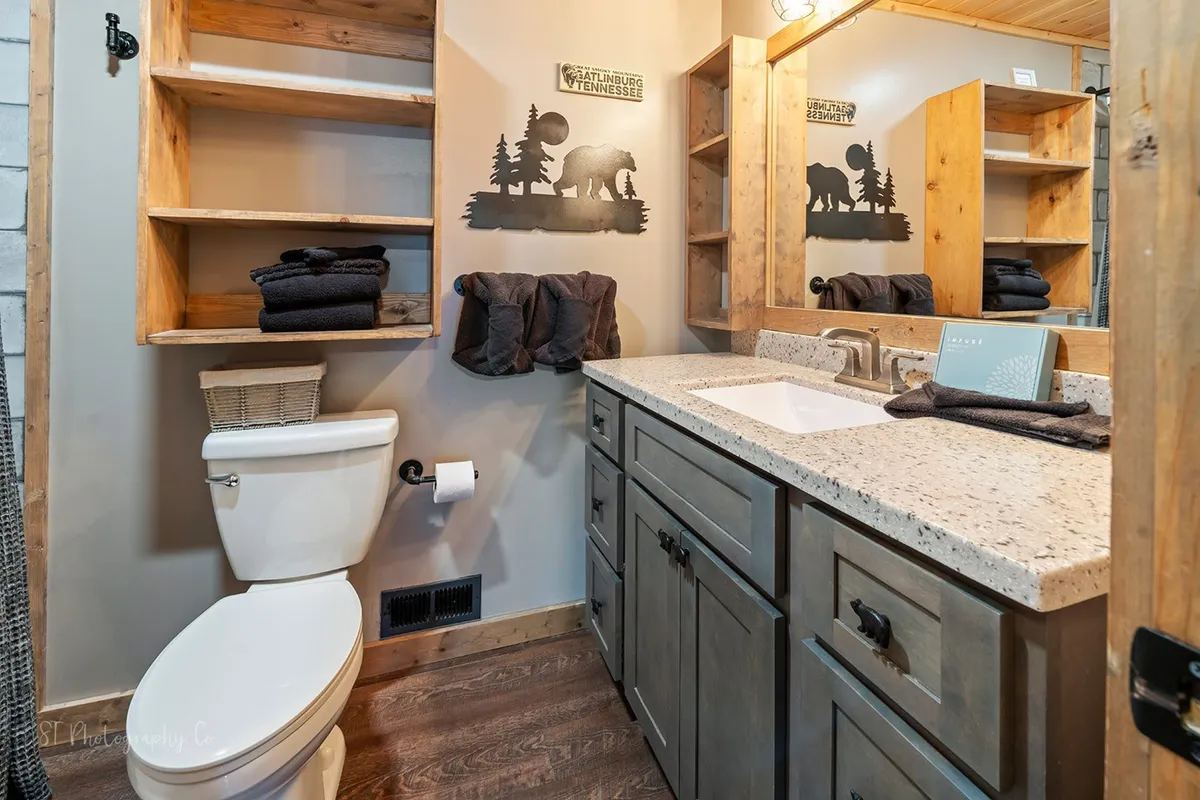 A bathroom scene with a wooden vanity, sink, shelves stacked with towels, and art featuring a bear silhouette. The space is accented with warm wooden details and soft lighting, creating a serene, inviting atmosphere for morning routines or relaxing baths.