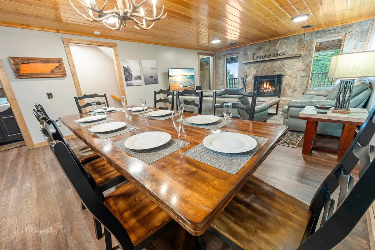 A warm, welcoming dining area anchored by a statement antler-style chandelier, a large wooden table set for eight, and a fireplace in the background. The space exudes rustic-chic charm with ample natural light from large windows, highlighting the communal and celebratory feel of the space.