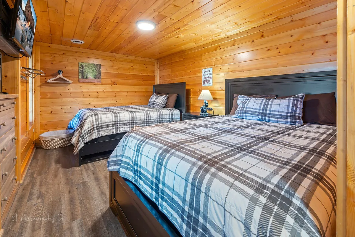 A downstairs bedroom with two queen beds dressed in classic plaid bedding, wooden walls that add rustic charm, a nightstand with lamp, and a wall-mounted TV for nighttime relaxation. The mood is relaxed and welcoming, ideal for families or groups seeking comfort and entertainment.