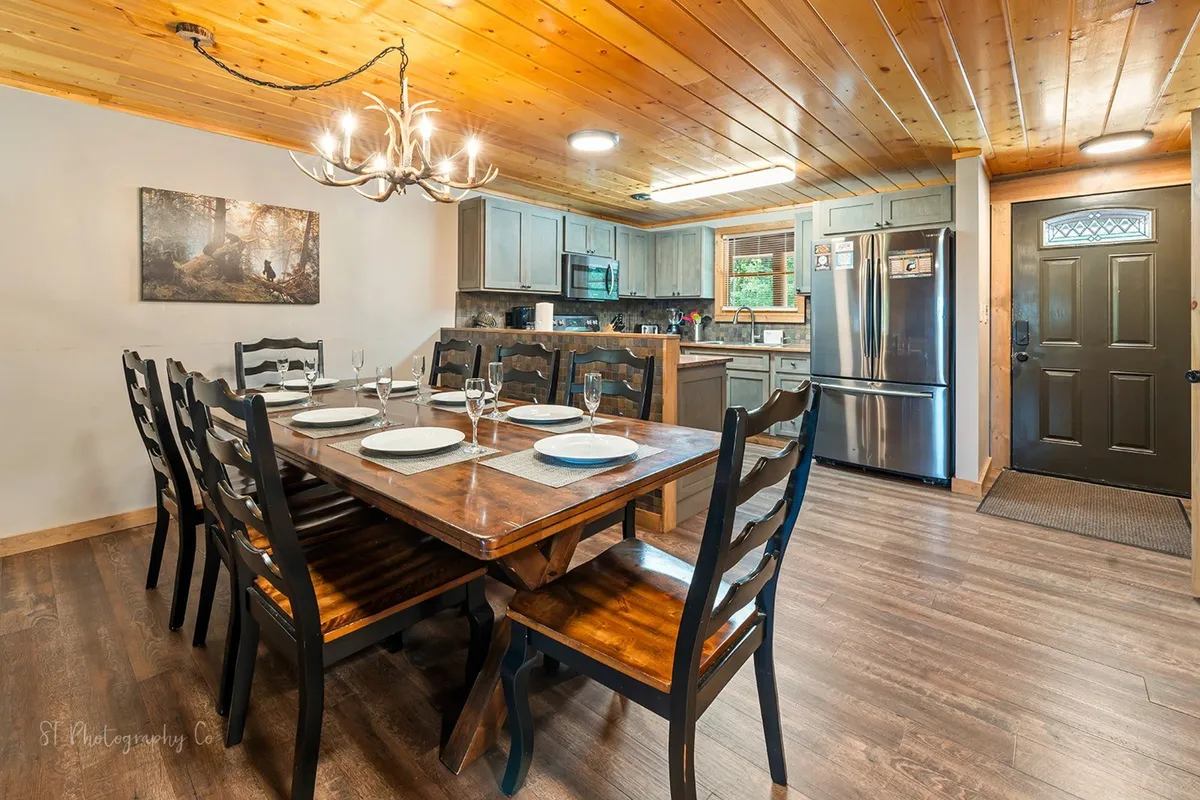 A bright, communal dining area with a large wooden table beneath a striking chandelier, modern kitchen appliances visible in the background, and art adorning the wall. The setup invites group meals and lingering conversations, making it ideal for family gatherings and celebrations.