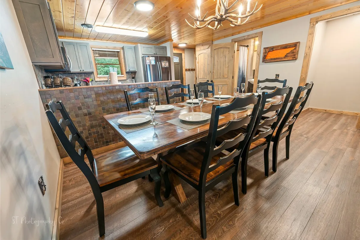 A warm and inviting dining room scene with a sturdy table set for a family meal, a soft chandelier overhead, and rich kitchen cabinetry behind. The ambiance suggests festive gatherings and intimate dinners alike.