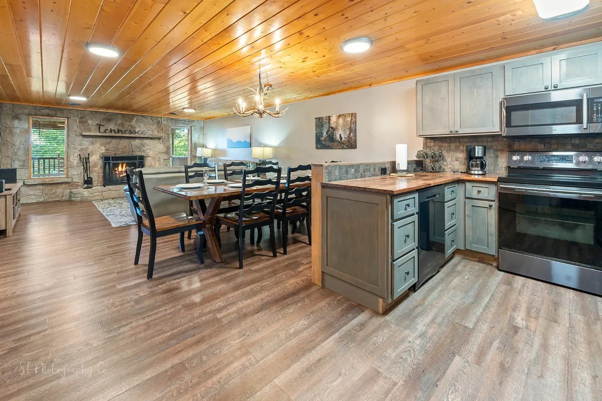 This image captures the open-plan living, dining, and kitchen area featuring a spacious dining table, a warm stone fireplace, a chandelier, and inviting rustic decor elements. The view lets you see how the spaces flow into one another, creating a communal atmosphere perfect for gatherings, meals, and game nights.
